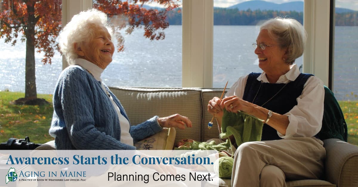 Two older women laughing together while one knits beside a window overlooking a lake
